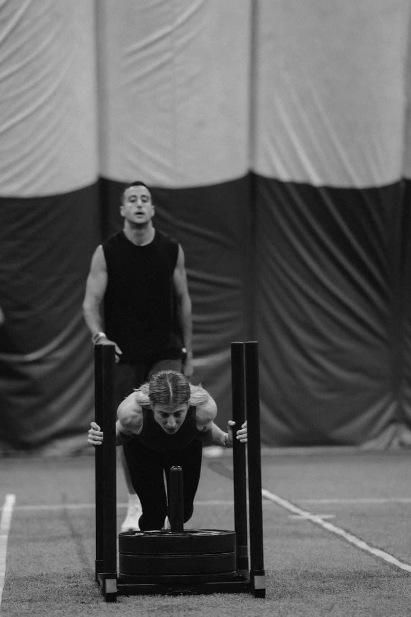 Black and white photo of my sister driving a loaded sled down indoor turf, head down and arms locked out, with me watching from behind