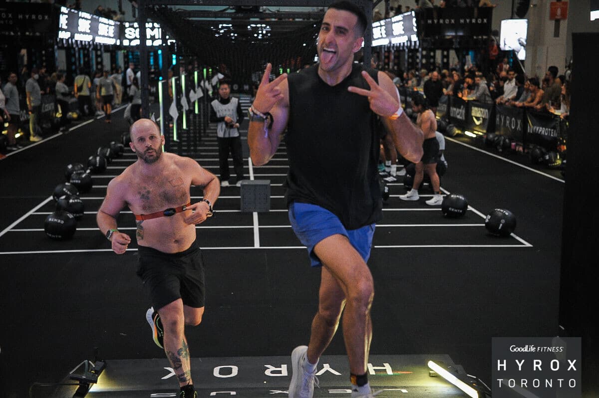 Running a lap at HYROX Toronto in a black vest and blue shorts, tongue out, throwing devil horns at the camera with the kettlebell floor and crowd behind
