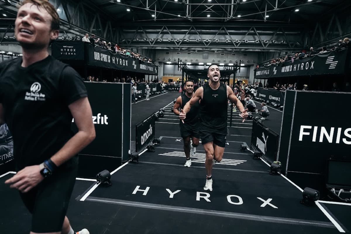 Sprinting down the finish chute at HYROX Vienna under the arena lights, arms driving, with the HYROX branding on the floor and a packed balcony above