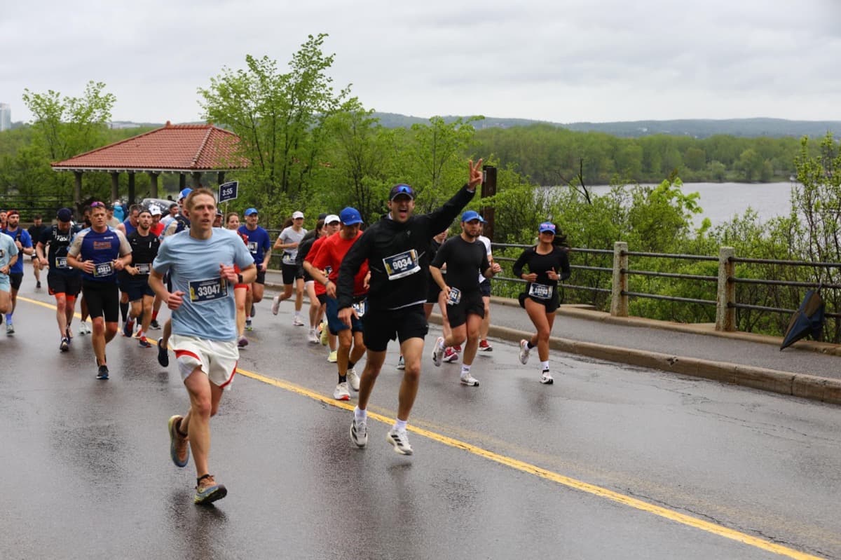 Mid-pack on a wet Ottawa Marathon course in a black jacket, one arm up in a peace sign, the river and the 3:55 pace bunny sign visible behind